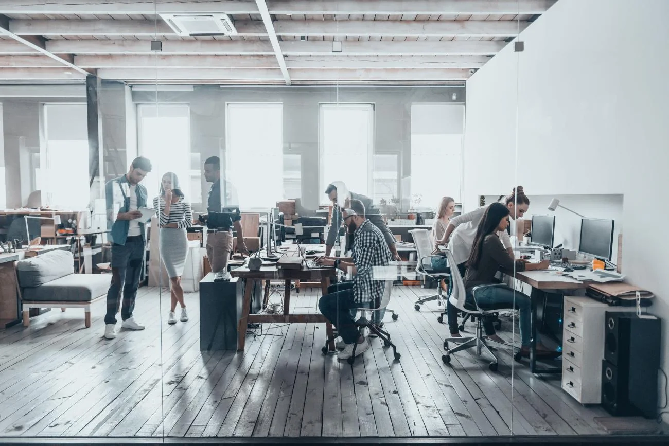 An open-plan office with teams working at desks behind a glass partition
